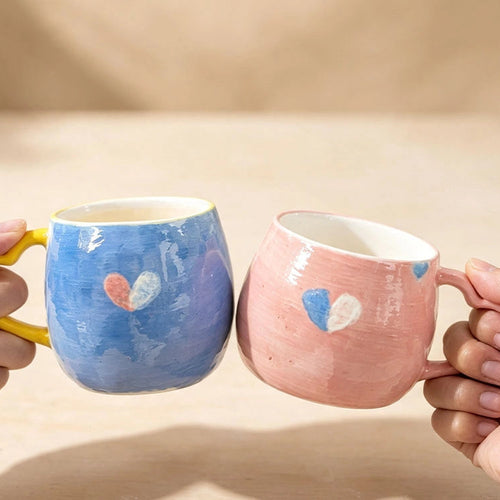 Two ceramic mugs held by hands in a toast-like pose. The left mug is light blue with a yellow handle and a small heart design in pink, white, and blue.