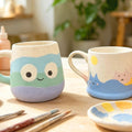 Two hand-painted ceramic mugs on a wooden table in a cozy craft studio setting. The mug on the left features a cute monster face design with pastel blue, mint green, and lavender sections.